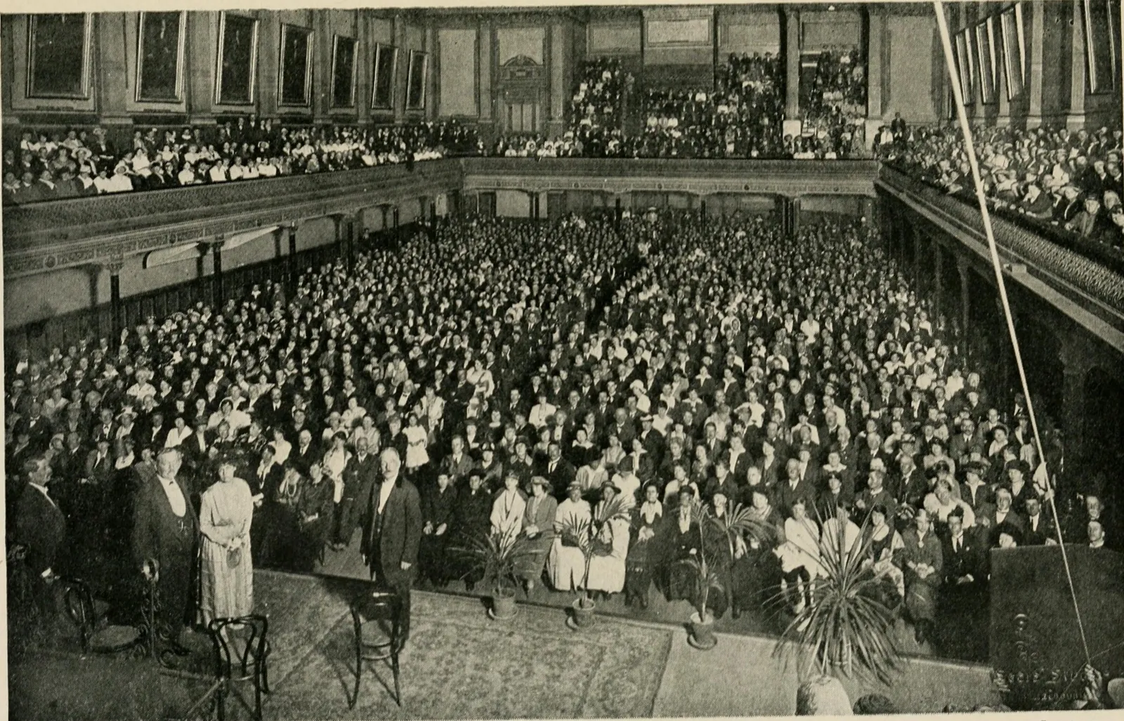 The Melbourne Town Hall seen from the platform, packed: thousands seated on the floor and in the galleries under the portraits on the walls, potted palms along the edge of the platform, and at the lower left a few people standing on the platform.