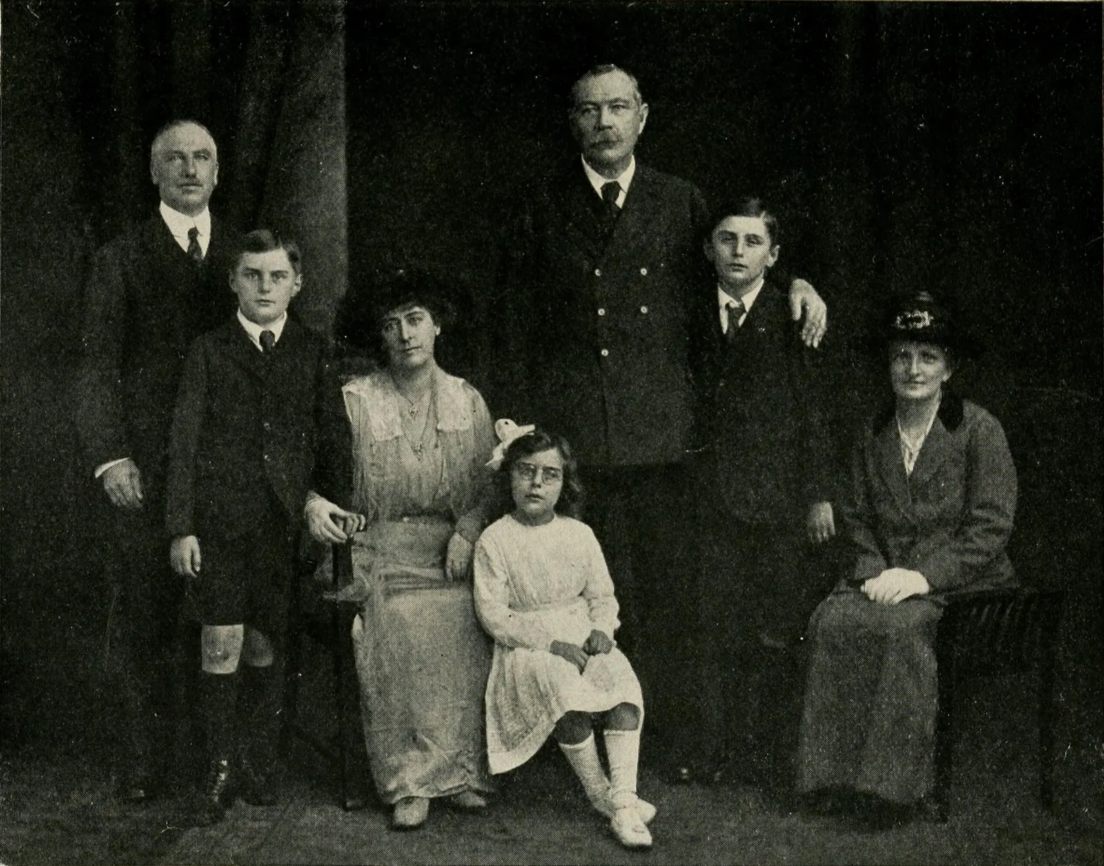 A studio group of seven against a dark curtain: Major Wood, standing, grey-haired; the two boys in dark suits; Lady Doyle seated in a pale lace dress with their small daughter; Conan Doyle; and Jakeman, the maid.