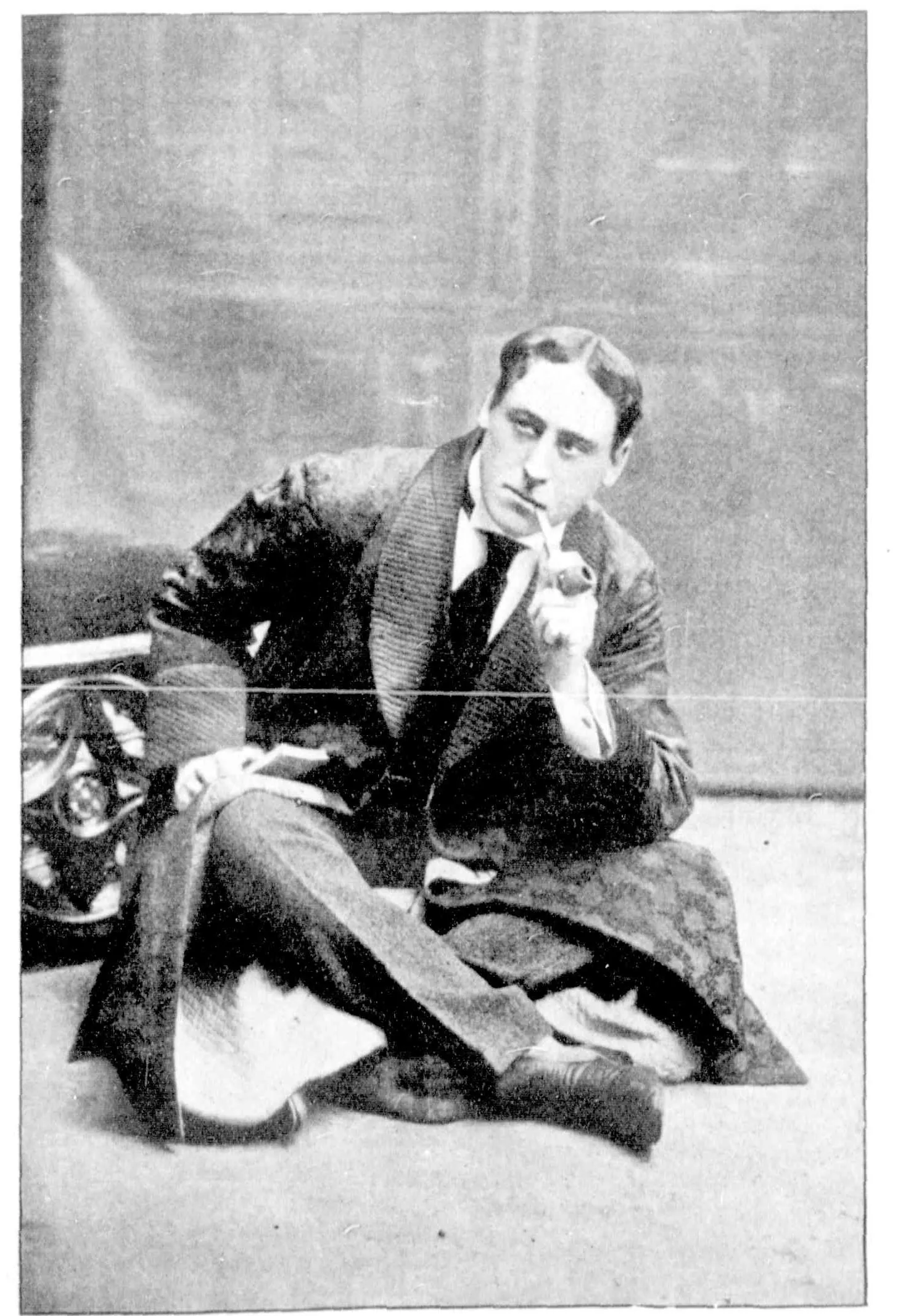 Cuyler Hastings as Holmes, sitting on the floor against a couch in a quilted dressing-gown over his suit, a curved pipe at his lips and a book on his knee.